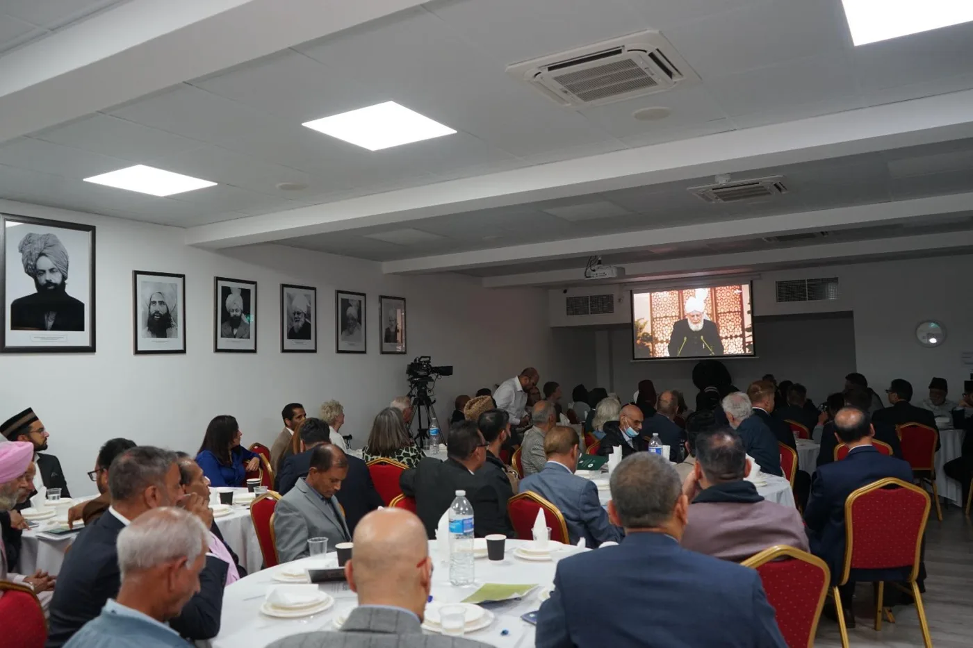Guests seated at round tables in a hall watching the keynote address on a large screen